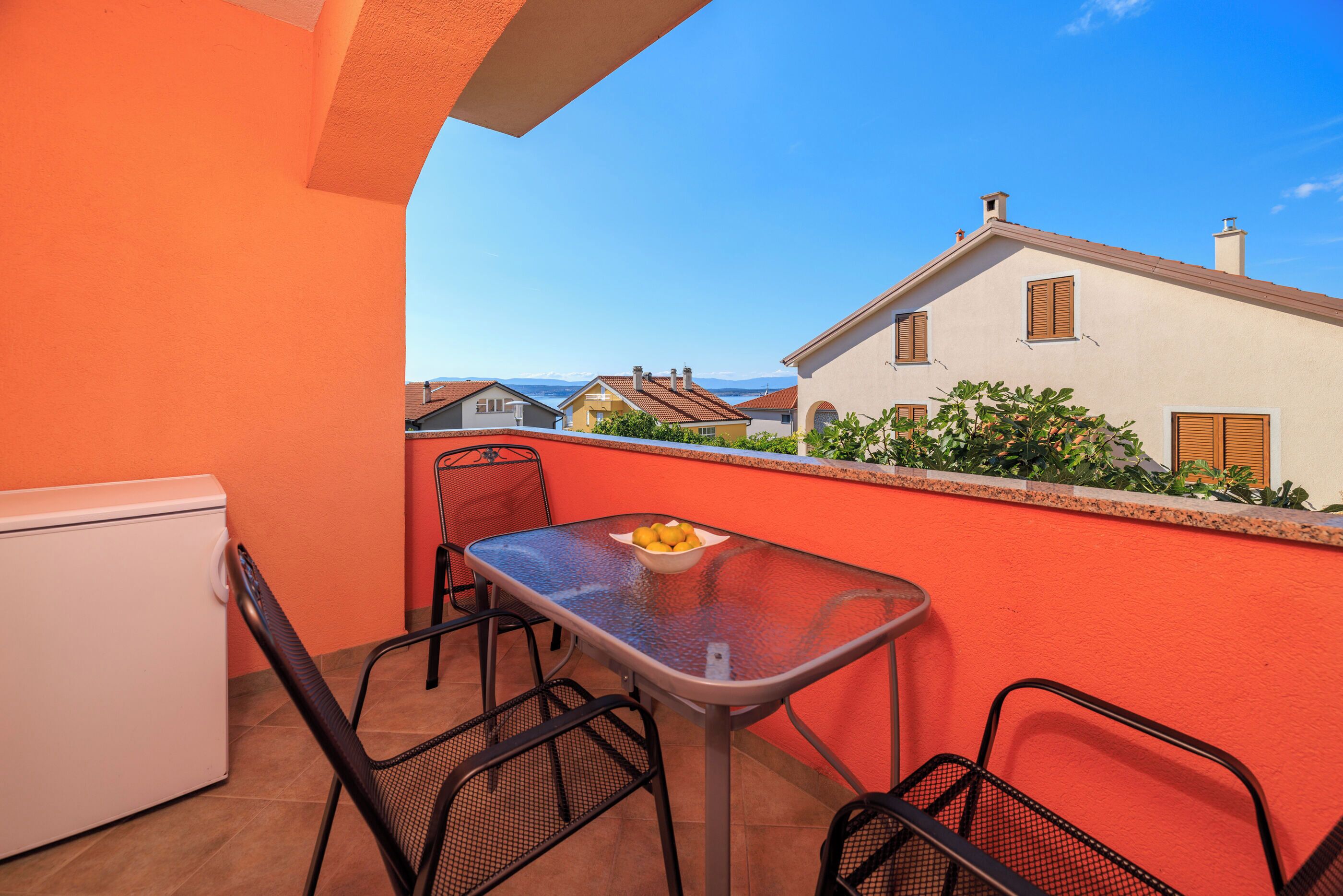 A balcony with a table and chairs, a bowl of fruit, and a view of houses in the distance.