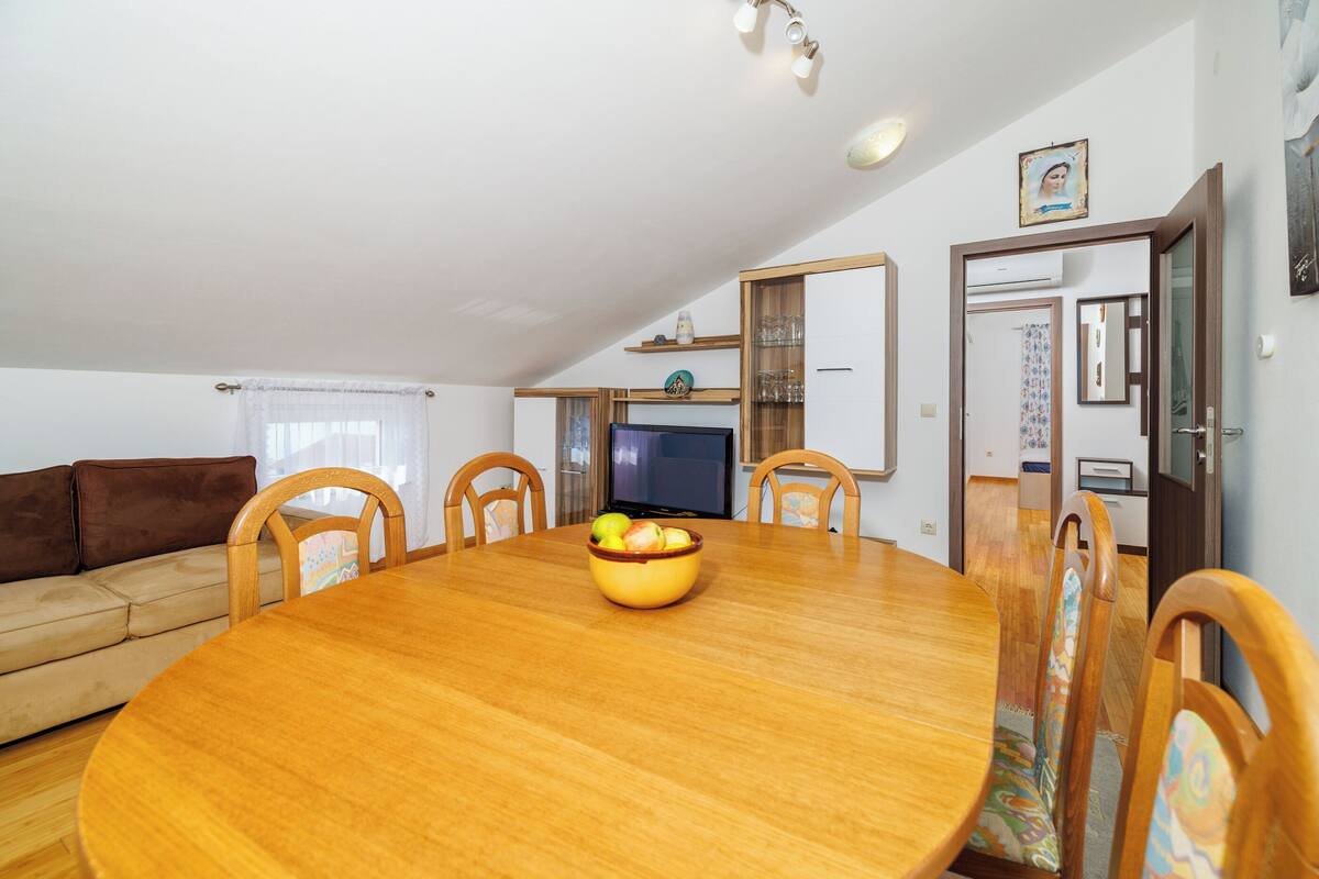 Ein Essbereich mit Holztisch, Stühlen und einer Obstschüssel. Im Hintergrund befindet sich eine Couch und ein Fernseher.