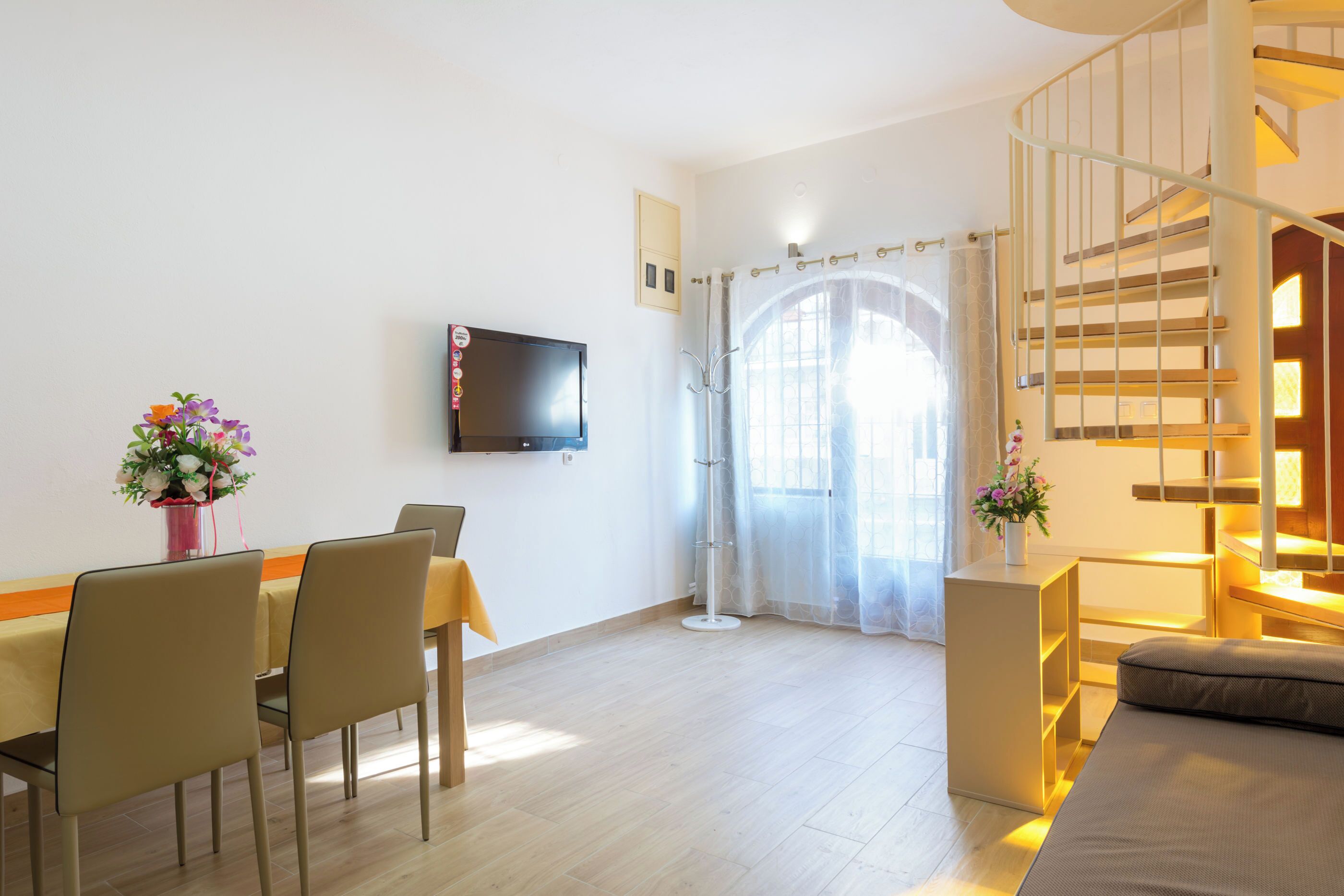 A modern living room with a dining area, a flat-screen TV mounted on the wall, a staircase, and a large window with sheer curtains.