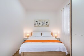 En pænt redt seng med hvidt og orange sengetøj, to sengeborde med lamper, og et blomsterbillede på væggen.