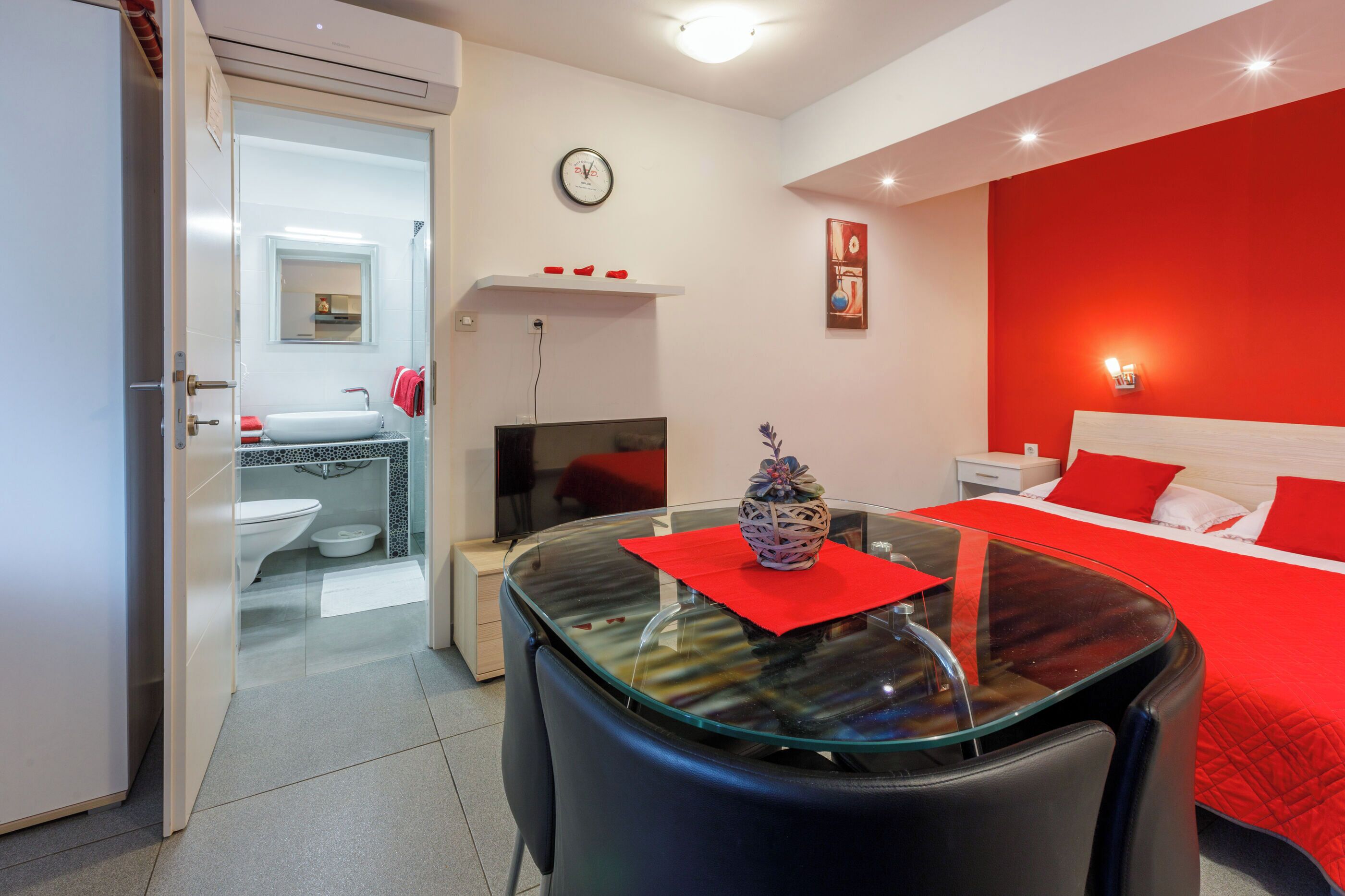A modern hotel room with a glass table, red accents, a bed with red bedding, a clock, and a small kitchen area.
