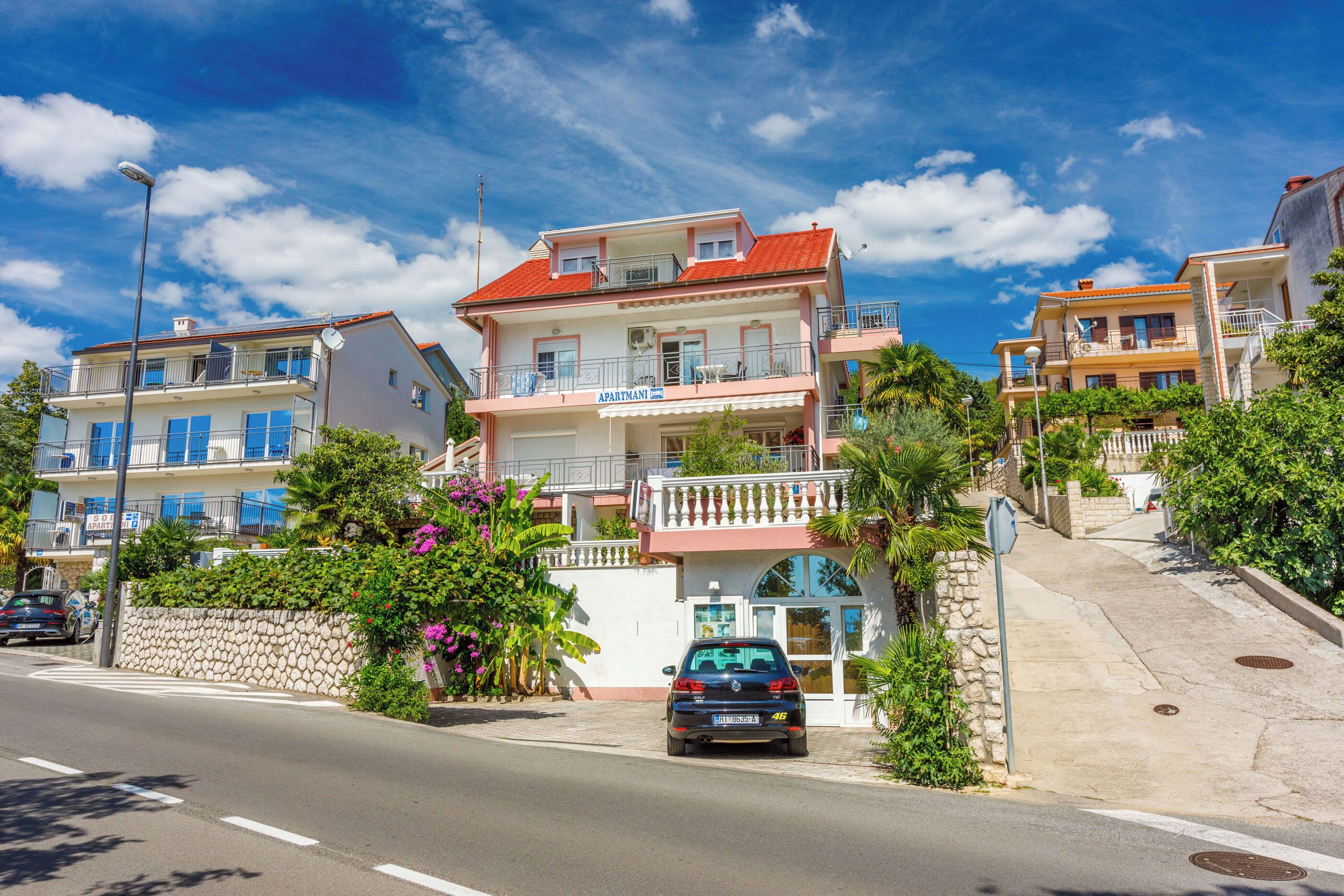 A multi-story residential building with balconies, a stone wall, and a car parked in front.