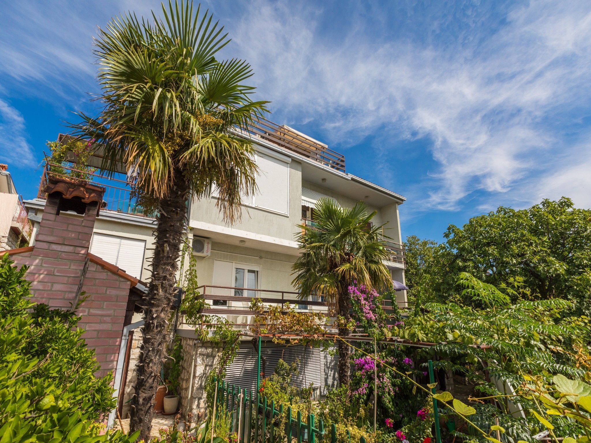 A two-story building with a balcony, surrounded by lush greenery and a clear blue sky.