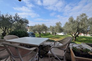 An outdoor patio with wicker chairs and a table, surrounded by olive trees and a view of a residential area. - Apartments Ljubomir (Cres)