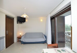 A room with a sofa, a television mounted on the wall, a table with a tablecloth, and a door leading to an outdoor area. - Apartments Ljubomir (Cres)