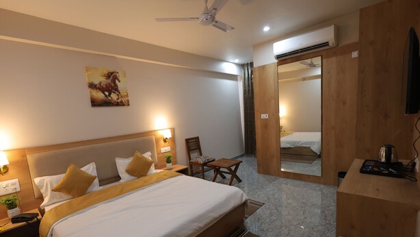 A hotel room with a large bed, a wooden headboard, a desk with a chair, a mirror, and a ceiling fan. - Royal Greens (Sikar)