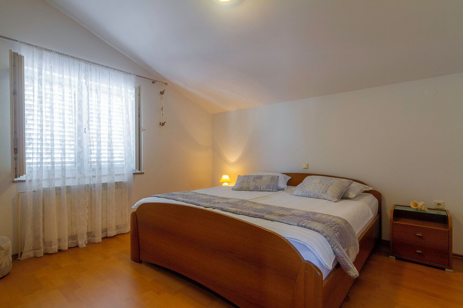 A bedroom with a wooden bed, a nightstand, a lamp, and a window with sheer curtains.