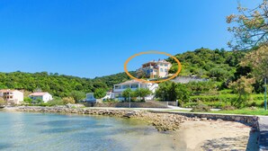 A coastal property with a clear view of the sea, a stone pathway leading to the beach, and a hillside with lush greenery. - Apartments Katica (Rab)