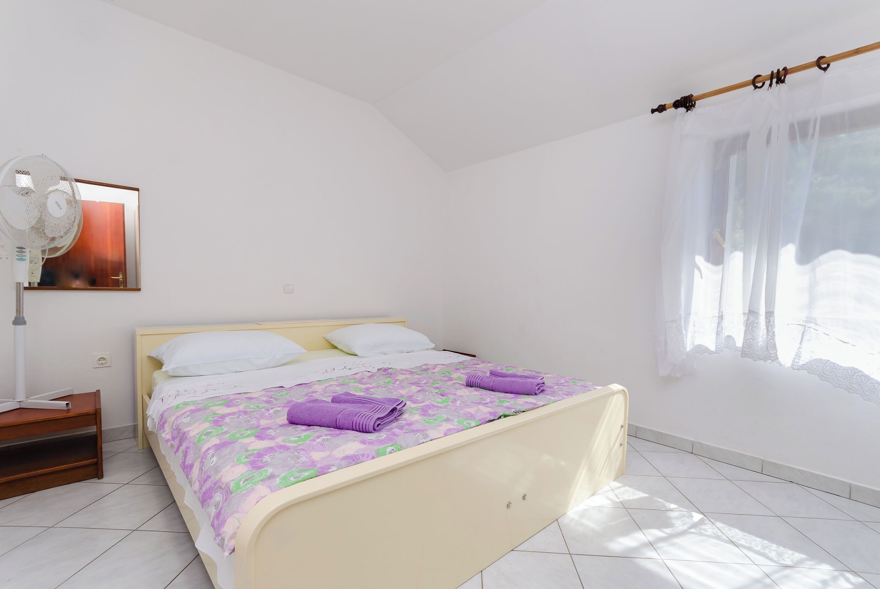 A bedroom with a bed, a nightstand, a fan, a window with sheer curtains, and a painting on the wall.