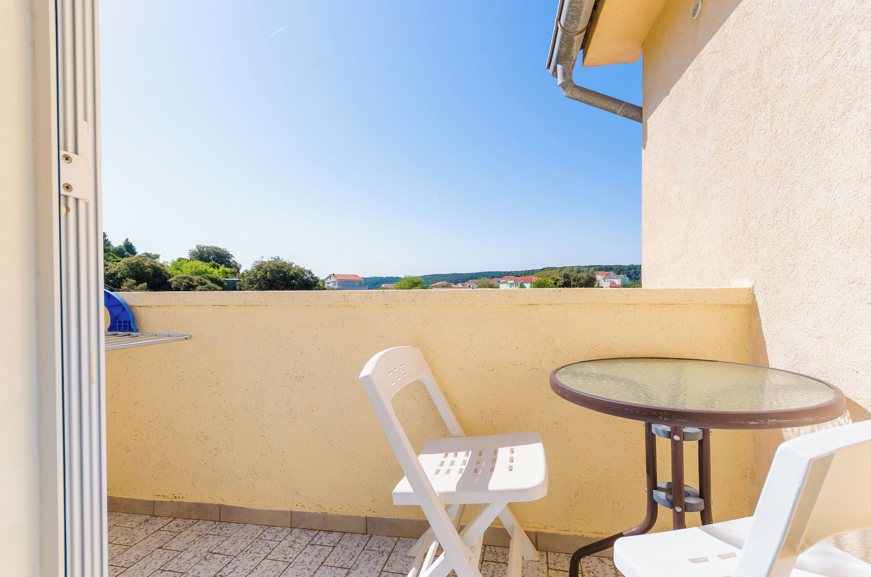 A balcony with a glass table and two white chairs, overlooking a distant landscape with houses and greenery.