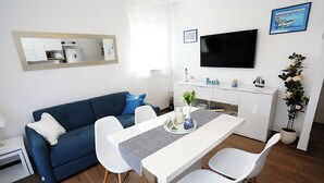A modern living room with a blue sofa, white dining table, and chairs. A flat-screen TV is mounted on the wall above the table. - Apartment Romarin (Supetar)