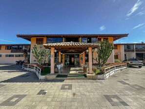 A building with a covered entrance, outdoor seating area, and parked vehicles. - Condomínio Portal do Atlântico (Porto Seguro)
