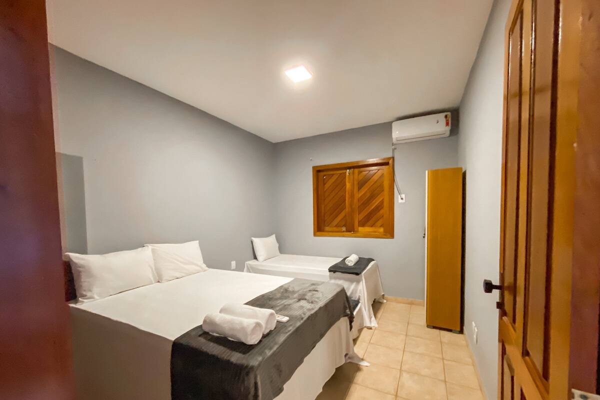 A hotel room with two beds, a small table, a window with wooden shutters, and an air conditioning unit on the wall.