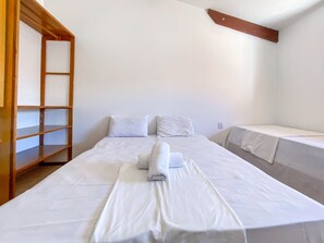 A neatly made bed with white linens, a wooden shelf unit, and a small bedside table. - Condomínio Portal do Atlântico (Porto Seguro)