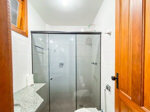 A modern bathroom with a glass-enclosed shower, white tiled walls, and a wooden door. - Condomínio Portal do Atlântico (Porto Seguro)