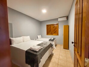 A hotel room with two beds, a small table, a window with wooden shutters, and an air conditioning unit on the wall. - Condomínio Portal do Atlântico (Porto Seguro)