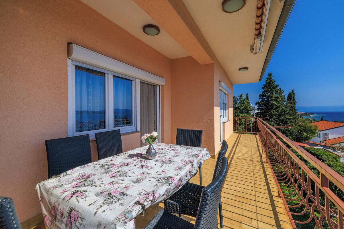 A balcony with a table and chairs, a railing, and a view of the sea and buildings.