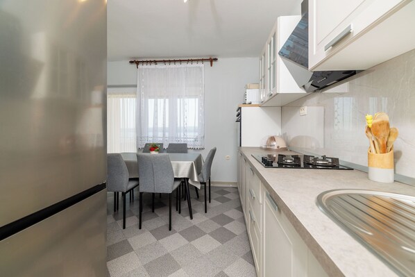 A modern kitchen with a dining area, featuring a checkered floor, a table with chairs, a stove, and a window with sheer curtains. - Apartments Melita (Crikvenica)