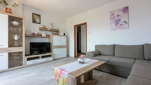 Uma sala de estar moderna com um sofá cinzento, uma mesa de centro de madeira e uma TV sobre um móvel de madeira. Há um armário branco com portas de vidro e uma prateleira acima.