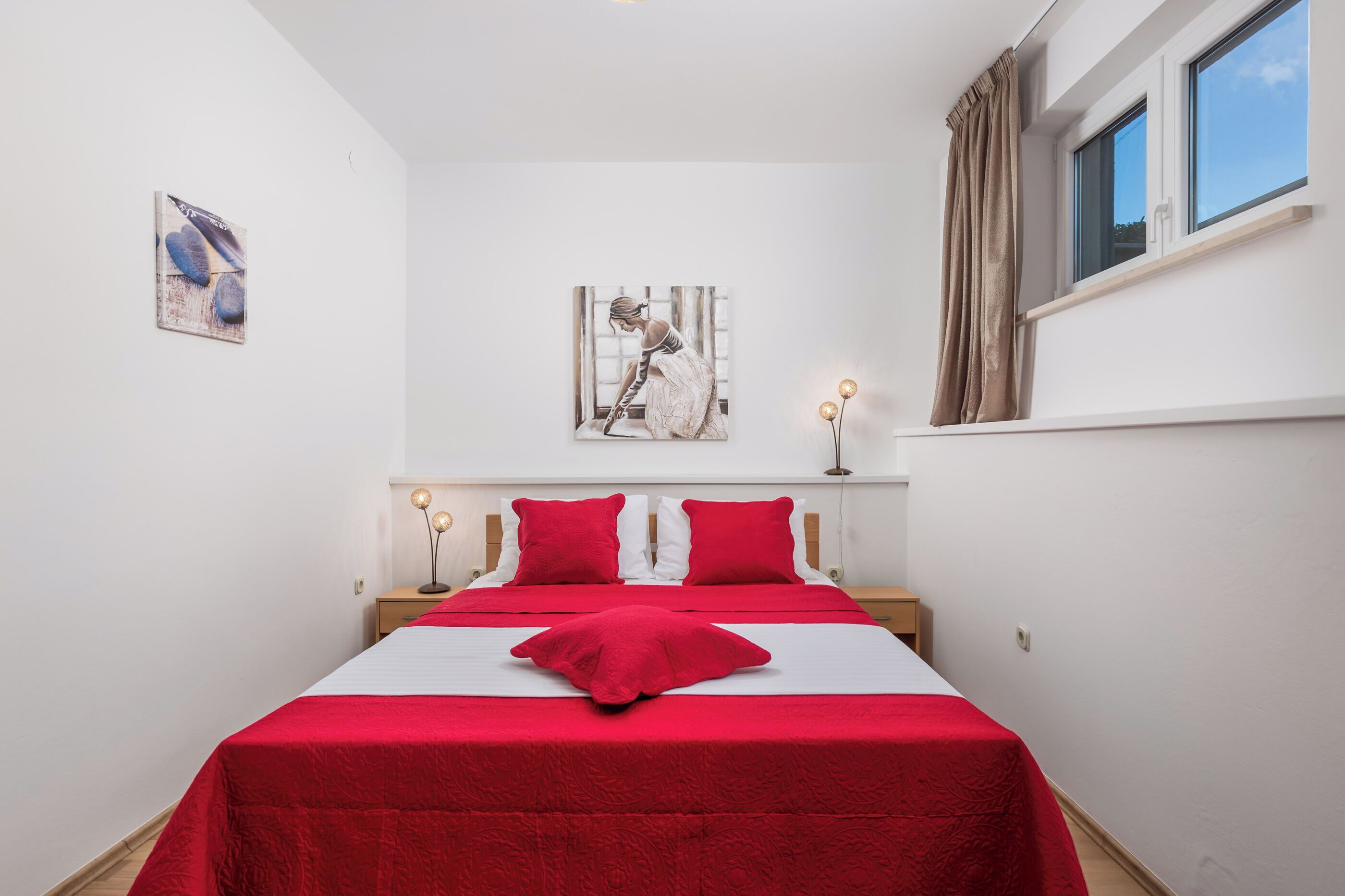 A bedroom with a large bed, red bedding, a painting on the wall, and a window with curtains.