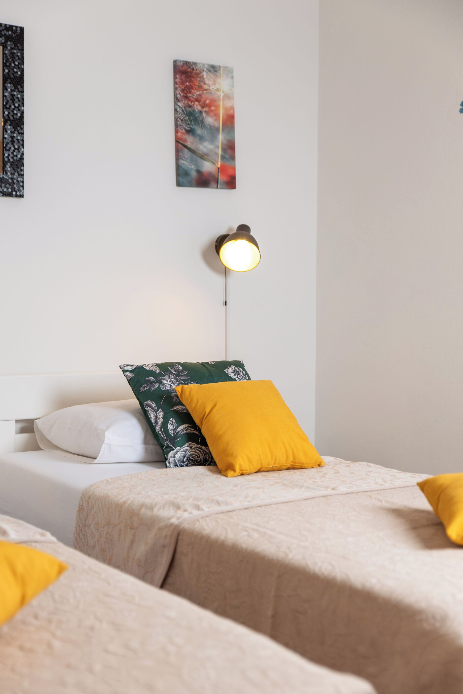 A hotel room with two beds, each with a patterned bedspread and a single yellow pillow. There is a wall-mounted lamp above the beds.