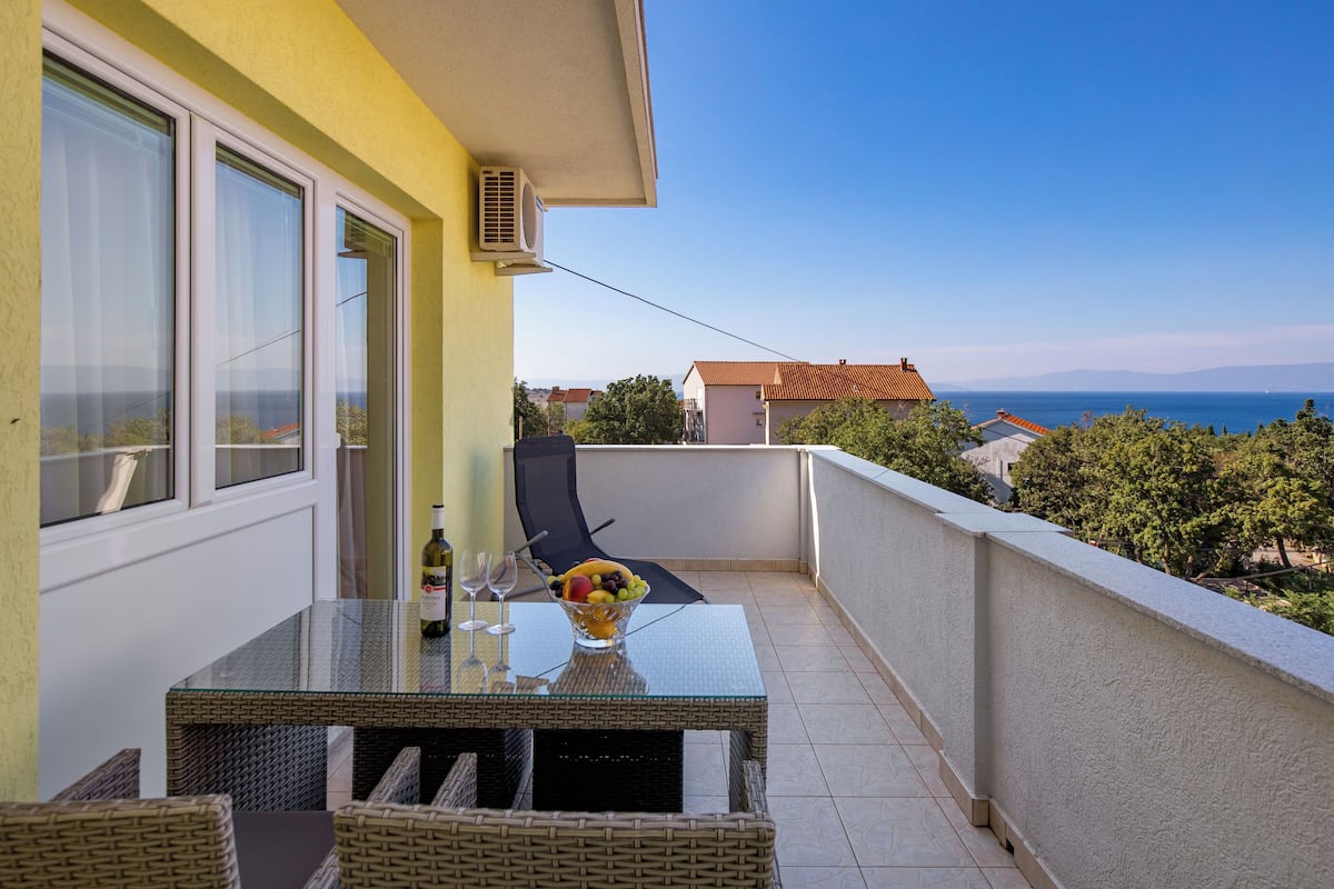 Un balcone con un tavolo in vetro, sedie in vimini e una ciotola di frutta.