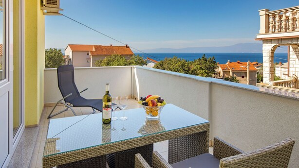 Un balcone con tavolo in vetro, sedie in vimini, una bottiglia di vino e una ciotola di frutta.