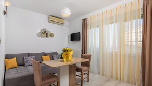 Un soggiorno moderno con un divano grigio, un tavolo da pranzo in legno e due sedie. Una TV a schermo piatto è montata a parete, e sul tavolo c'è un bouquet di fiori.