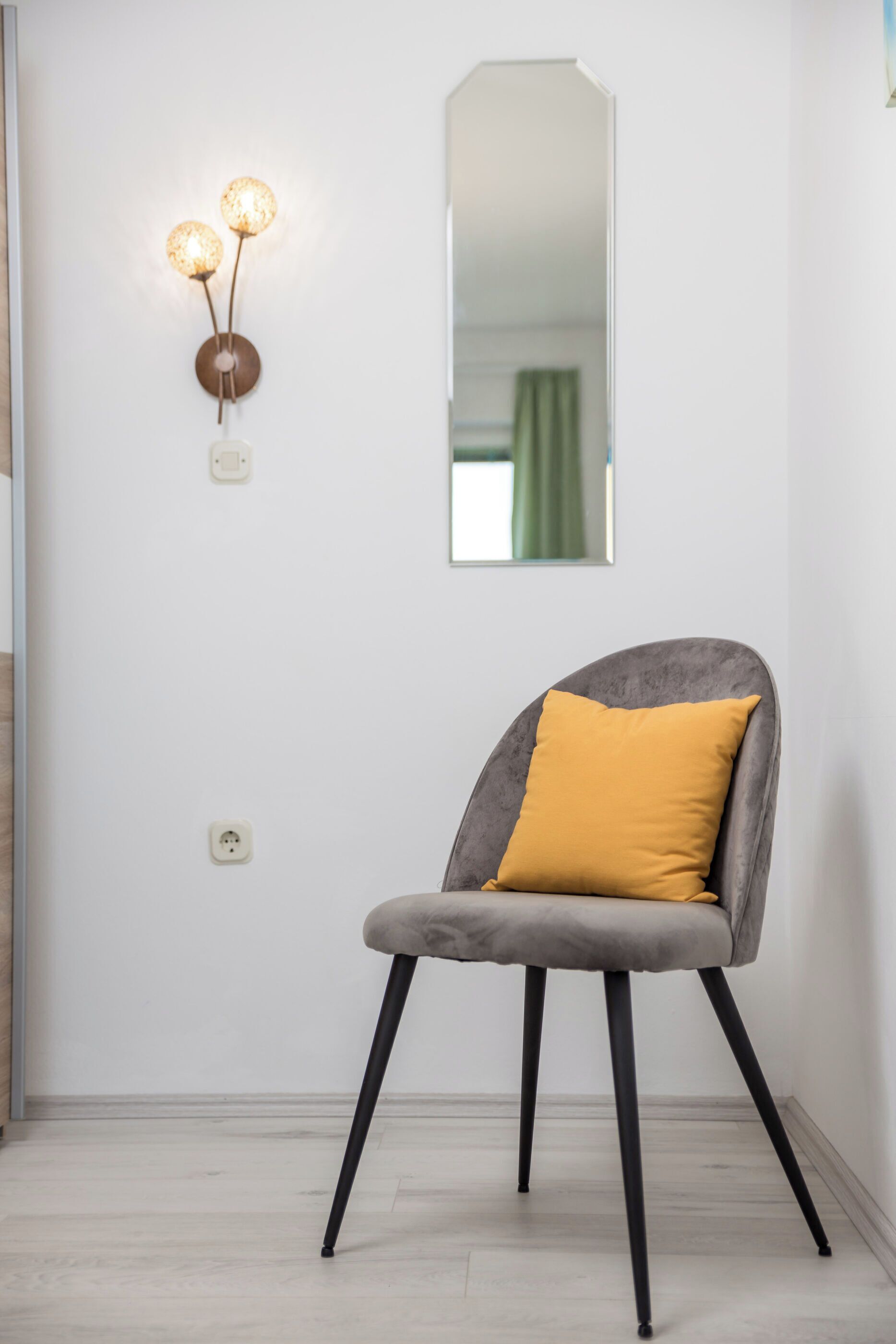 A chair with a mustard-colored cushion in a room with a white wall, a round mirror, and a wall-mounted light fixture.
