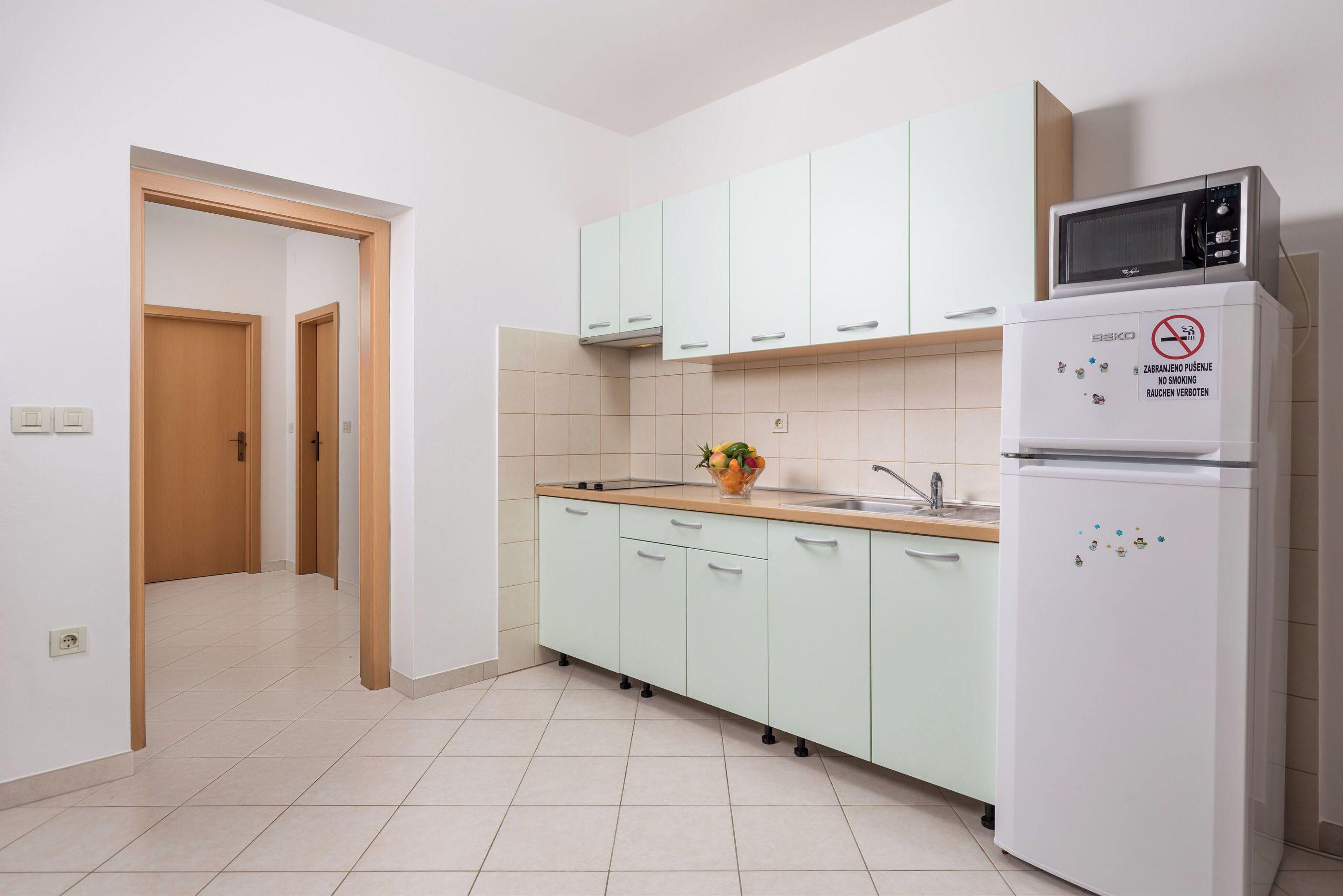 A kitchen with light green cabinets, a microwave, a refrigerator with a smoking sign, and a fruit bowl on the counter.