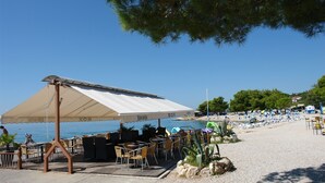 An outdoor seating area with tables and chairs under a large canopy, overlooking a beach with umbrellas and lounge chairs. - Apartments Matilda (Crikvenica)