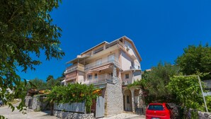 A multi-story building with balconies, a stone base, and a red car parked in front. - Apartments Matilda (Crikvenica)