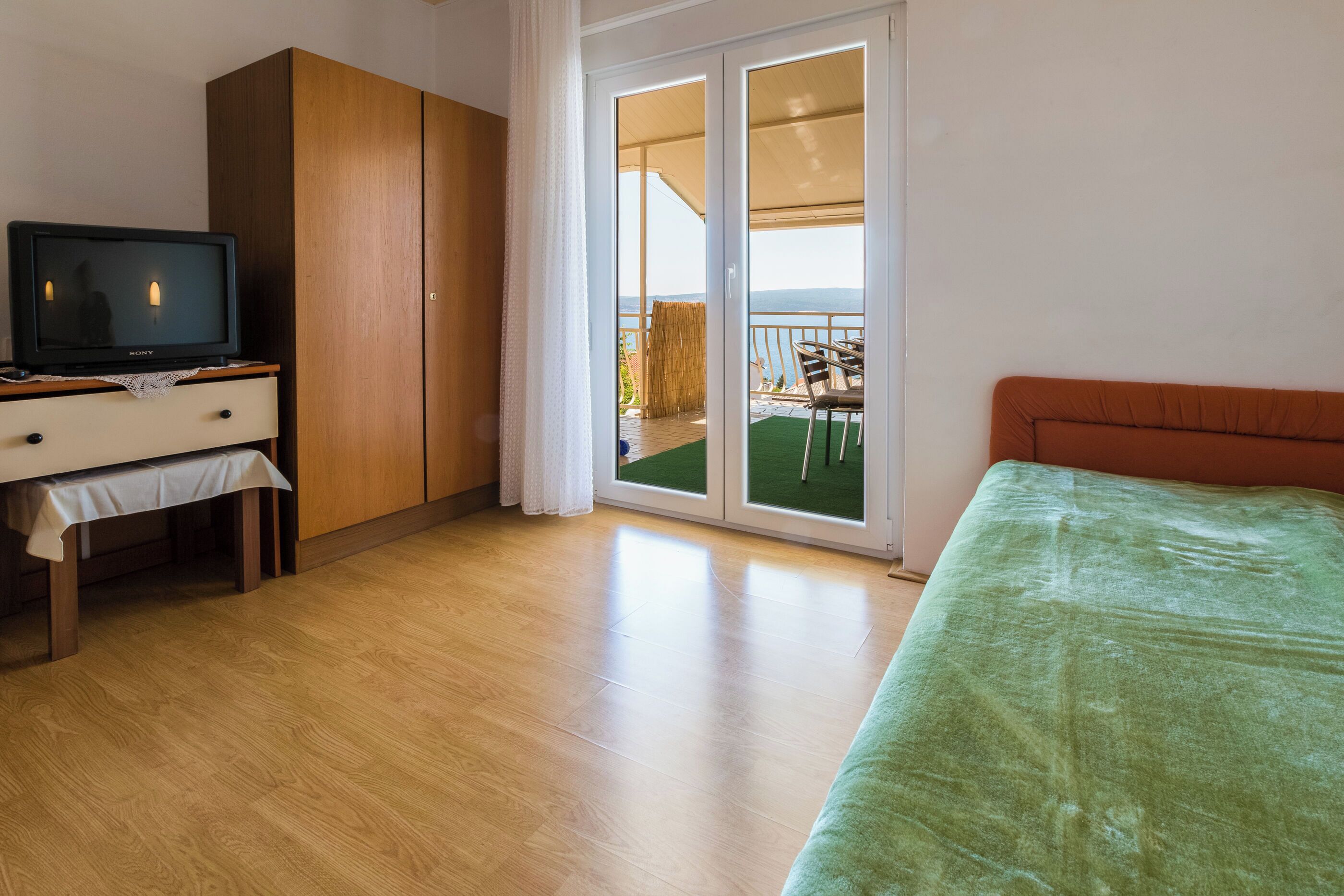 A room with a bed, a TV on a wooden cabinet, a sliding glass door to a balcony with outdoor furniture.