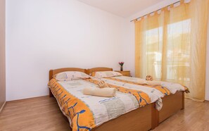 Un llit ben arropat amb un llençol estampat i dos coixins, una taula de nit amb un ram de flors, i una finestra amb una cortina transparent.