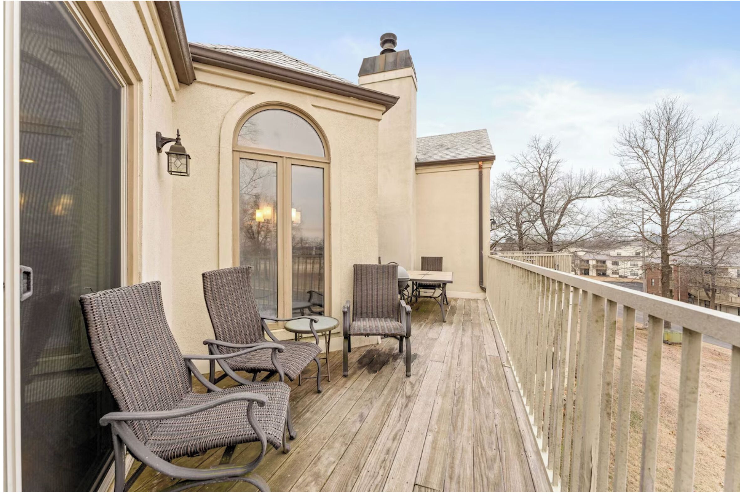 A covered outdoor area with wicker chairs and a table, a large arched window, and a view of trees and buildings.