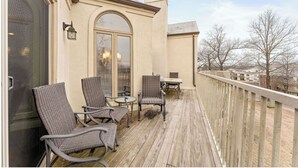 Ein überdachter Außenbereich mit Korbstühlen, einem Tisch, einem großen Bogenfenster und Blick auf Bäume und Gebäude.