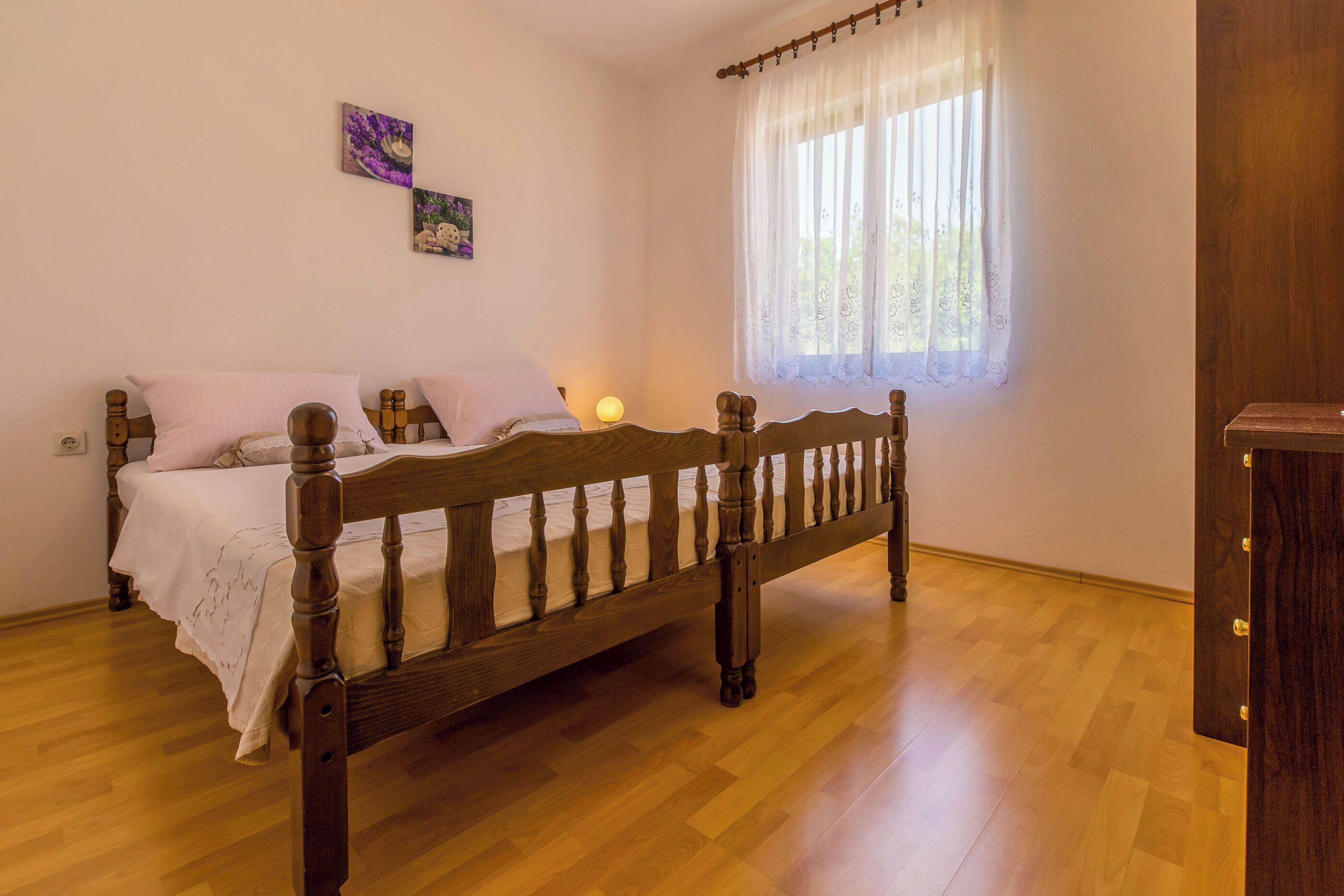 A bedroom with a wooden bed, white bedding, a bedside lamp, and a window with sheer curtains.