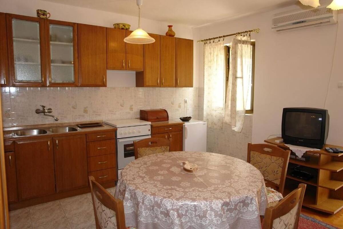 A compact kitchen with wooden cabinets, a stove, a refrigerator, a dining table with chairs, and a television.