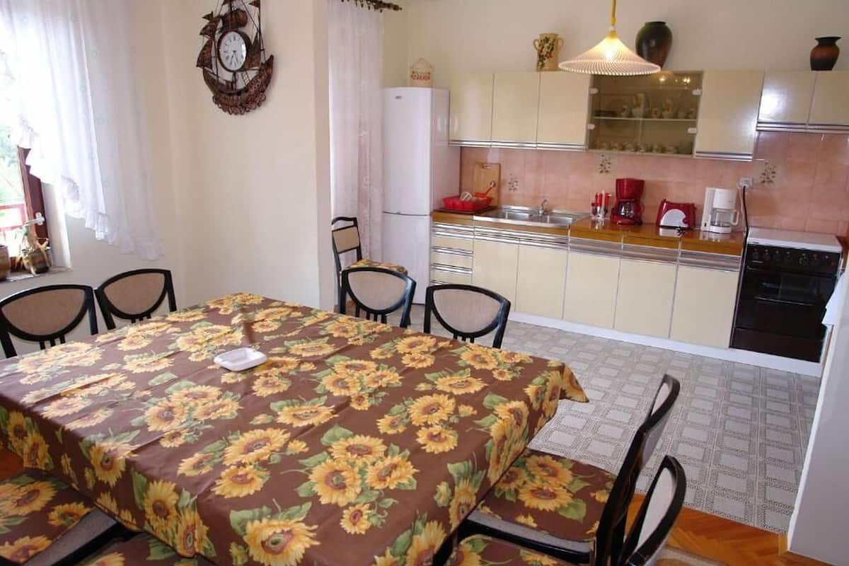 A dining area with a table covered in a sunflower-patterned tablecloth, a kitchen with a refrigerator, stove, and various kitchen items.