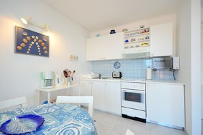 Une cuisine compacte avec des meubles blancs, un four à micro-ondes, une bouilloire et une machine à café. On y trouve une table à manger recouverte d’une nappe bleue et agrémentée d’une assiette décorative.
