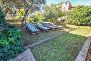 Ein Garten mit vier Liegestühlen und einem kleinen Tisch, umgeben von viel Grün und einem Gebäude im Hintergrund.