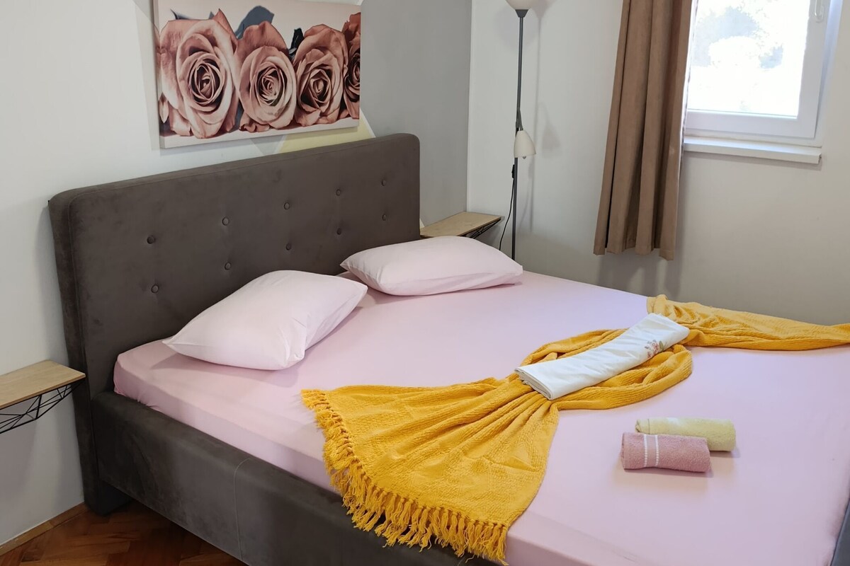 A bedroom with a bed, pillows, a blanket, a painting, a floor lamp, and a window with curtains.