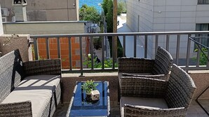 Un balcón con mobiliario de mimbre y una pequeña mesa, con vistas al mar y a una calle con edificios.