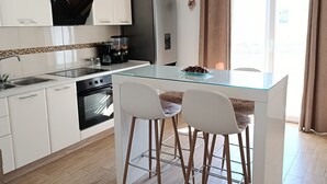 Una cocina moderna con armarios blancos, un refrigerador de acero inoxidable, un horno empotrado y una mesa de comedor de vidrio con dos taburetes altos.