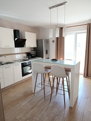 A modern kitchen with white cabinets, a stainless steel refrigerator, a built-in oven, and a glass dining table with two bar stools. - Apartments Lea-stella (Zadar)