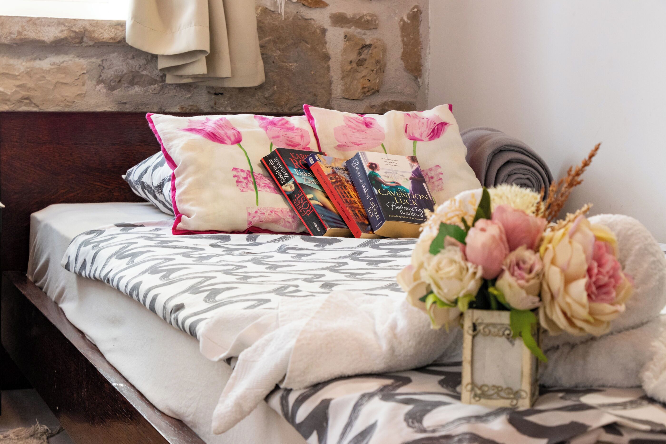 A neatly made bed with a patterned bedspread, pillows with floral designs, a stack of books, and a vase with flowers.