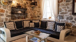 A cozy living room with a stone wall, a sofa with patterned cushions, a glass coffee table, and a window with curtains. - House Ston (Orebic)