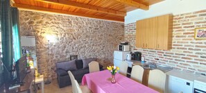 Una stanza accogliente con parete in pietra, soffitto in legno, un divano, un tavolo da pranzo con tovaglia rosa e un angolo cottura con forno a microonde e pensili.