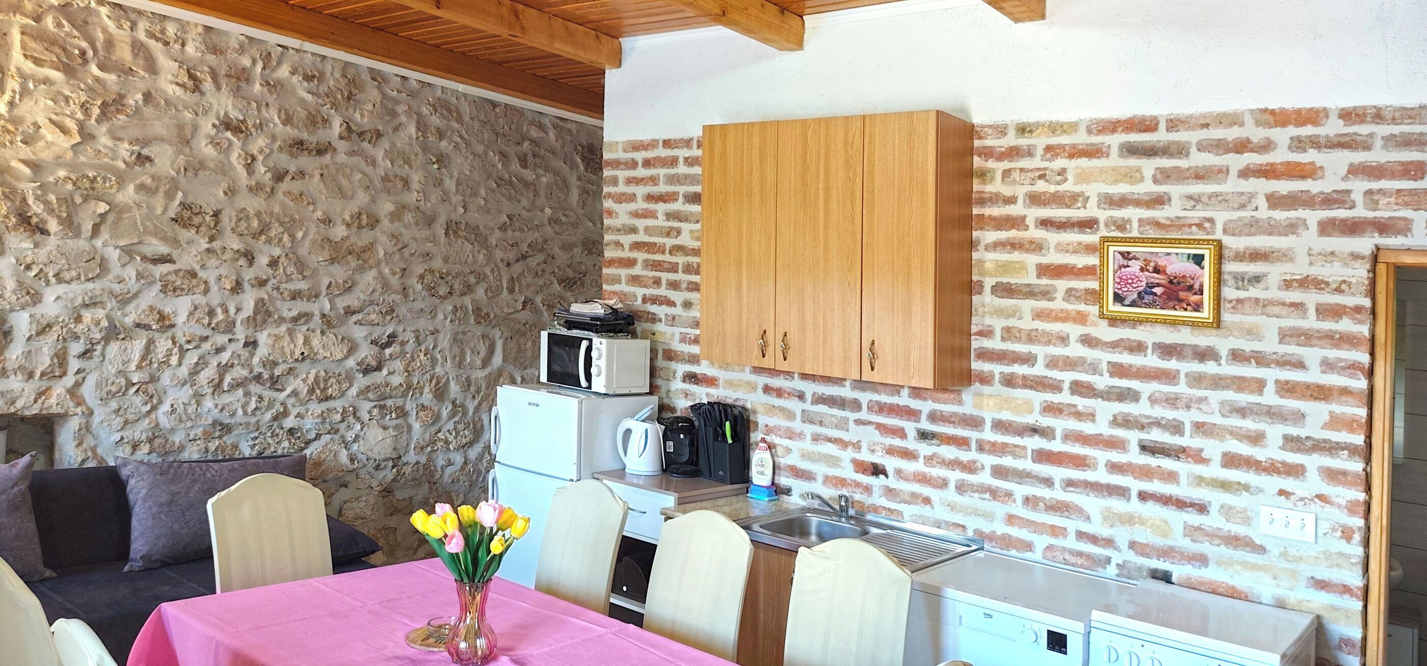 A room with a stone wall, wooden ceiling, a kitchen area with appliances, and a dining table with chairs.