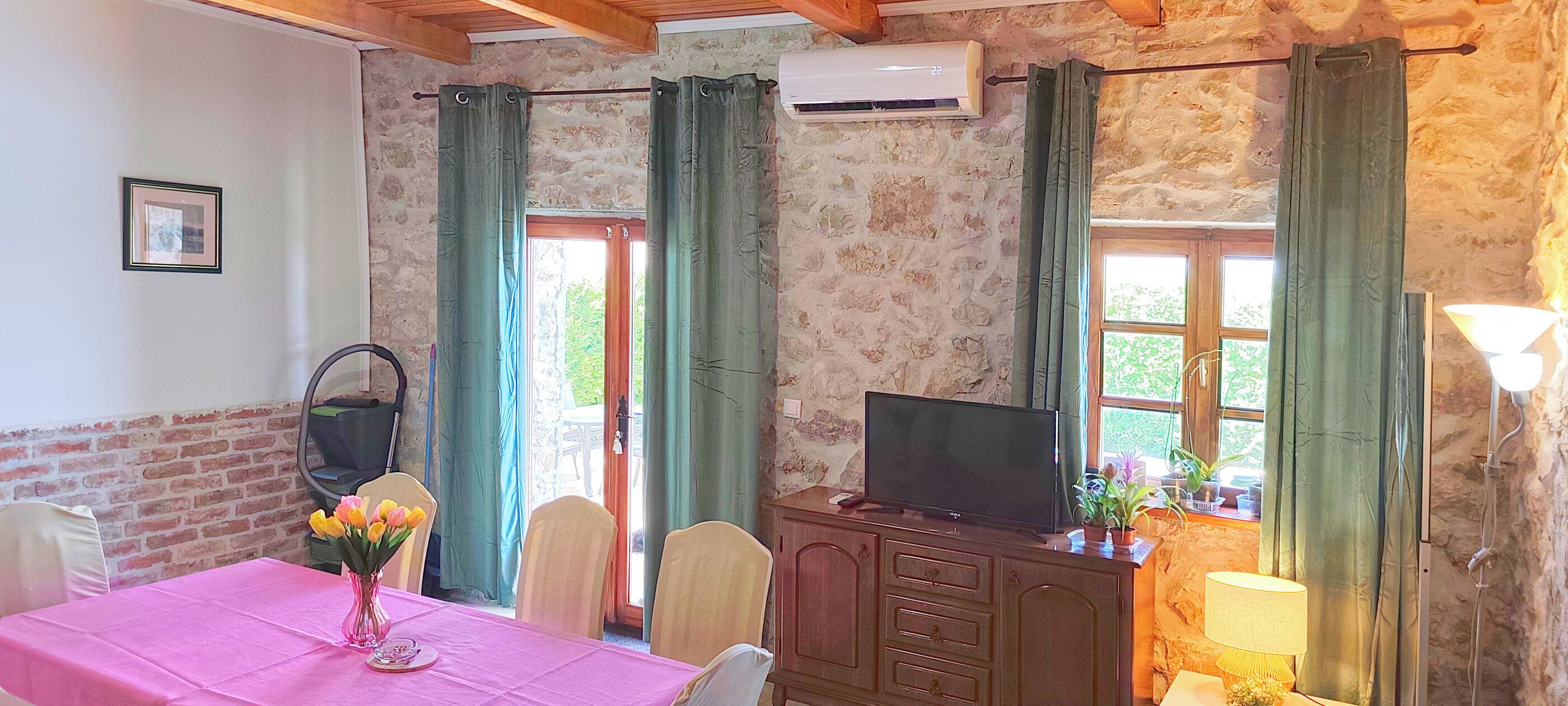 A room with a brick wall, a TV on a wooden cabinet, a table with a pink tablecloth, chairs, and a vase of flowers.